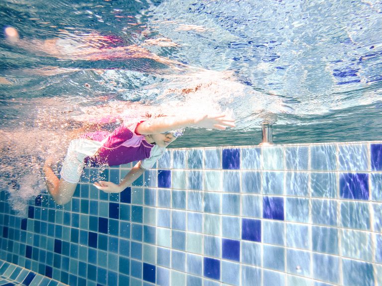 Underwater young little cute girl is swimming in the swimming pool with her swimming teacher. Seen under water while she is diving forward ahead of her. Swimming kid in the pool concept.